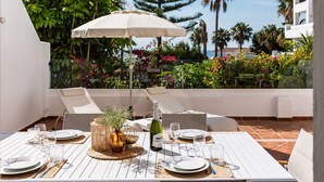 A sunlit outdoor dining area with a table set for four, surrounded by white chairs, a large umbrella, and lush greenery in the background.