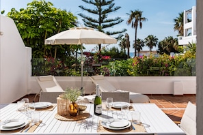 A sunlit outdoor dining area with a table set for four, surrounded by white chairs, a large umbrella, and lush greenery in the background.