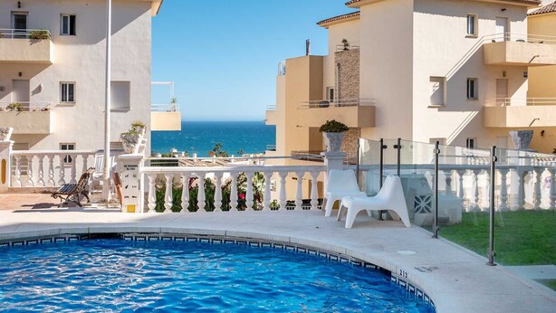 A swimming pool with a view of the sea, surrounded by white chairs and a white fence.