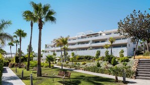 A modern hotel with multiple floors, a well-maintained garden with palm trees, and a clear sky. - Olas Sunset by Alfresco Stays (Estepona)