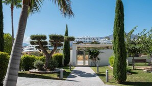A pathway leading to a modern building with a glass entrance, surrounded by palm trees and cypress trees, with a mountain in the background. - Olas Sunset by Alfresco Stays (Estepona)