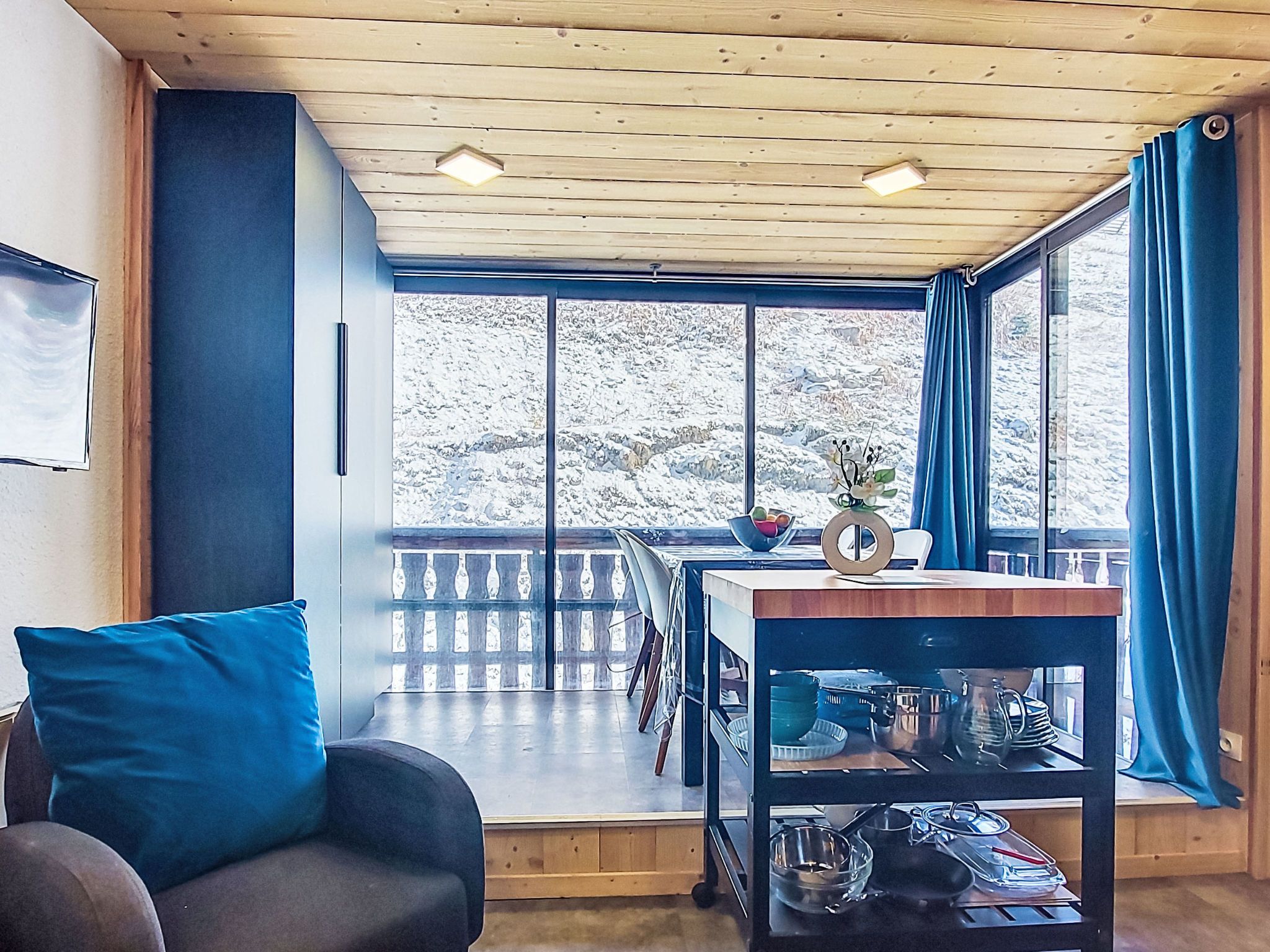 Une pièce avec un plafond en bois, un canapé agrémenté d’un coussin bleu, une table à manger avec des chaises, et une vue sur un paysage enneigé par de grandes fenêtres.