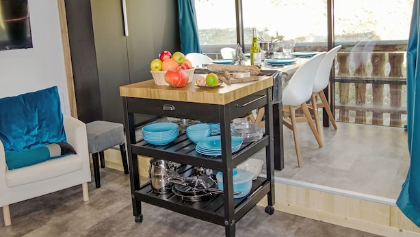 A modern kitchen with a wooden table set for dining, a fruit basket, and a wine bottle on the counter.