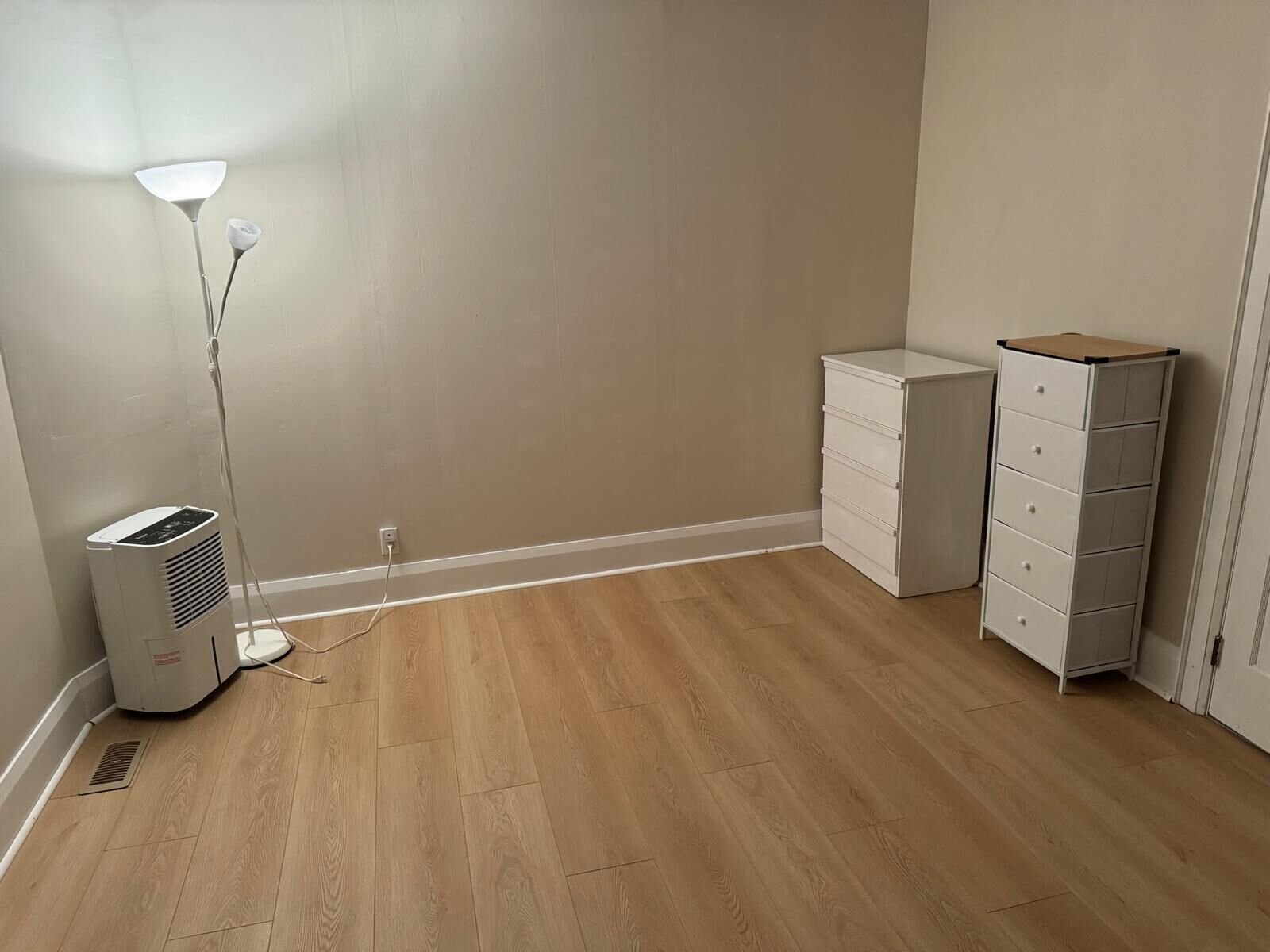 Una habitación con piso de madera, un calentador portátil, un aparador blanco y una lámpara de pie.