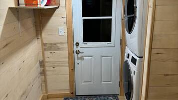 A laundry room with a washing machine, a door, and a shelf with cleaning supplies.
