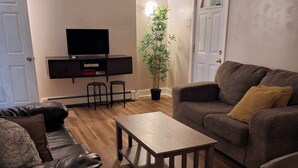 A living room with a leather sofa, a wooden coffee table, a grey sofa with a yellow pillow, a TV on a black stand, and a potted plant.