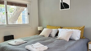 Una cama bien hecha con un edredón gris, almohadas blancas y una almohada decorativa amarilla. Se ve una ventana con persianas, una obra de arte colgada en la pared y un piso de madera.