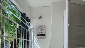 Una habitación de esquina con ventana, puerta y un cartel en la pared. Hay plantas en macetas sobre el suelo.
