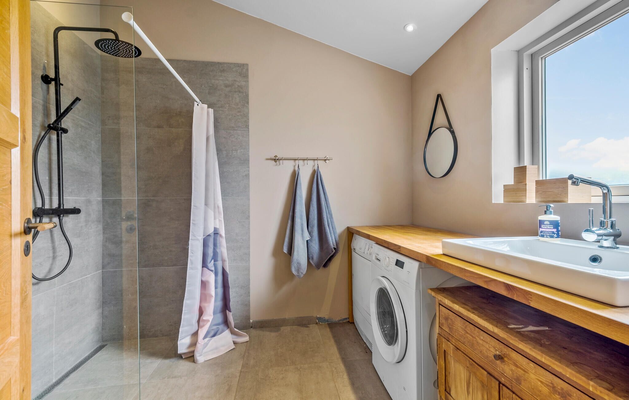 Un baño moderno con ducha de vidrio, lavadora, lavamanos y ventana con vistas.