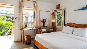 Une chambre à coucher avec un lit, une table de chevet en bois, une petite table sur laquelle est posé un vase de fleurs, et une vue sur l’extérieur.