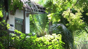 Un bâtiment blanc doté d’une fenêtre et d’une structure en bois surmontée d’un toit de chaume, le tout entouré d’un feuillage verdoyant et dense.