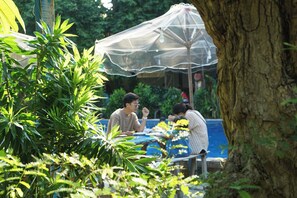 A dining area with tables and chairs, surrounded by lush greenery and a large tree trunk. - Stop And Go Lang Chai Bau Trang (Bac Binh)
