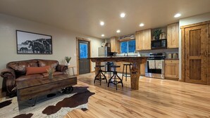 Un vaste salon comprenant un canapé en cuir, une table basse en bois et un tapis en peau de vache. À côté se trouve une cuisine avec des meubles en bois, un îlot central doté de tabourets et un réfrigérateur.