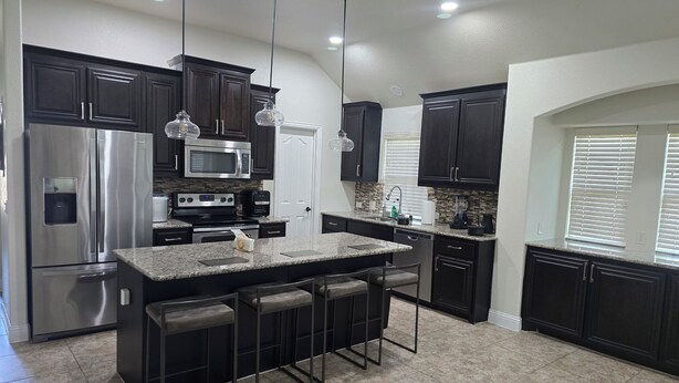 A modern kitchen with dark cabinets, stainless steel appliances, a granite countertop, and pendant lighting.