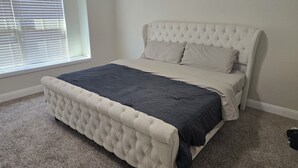 A bedroom with a tufted headboard, a bed with a dark and light-colored bedspread, and a window with blinds.