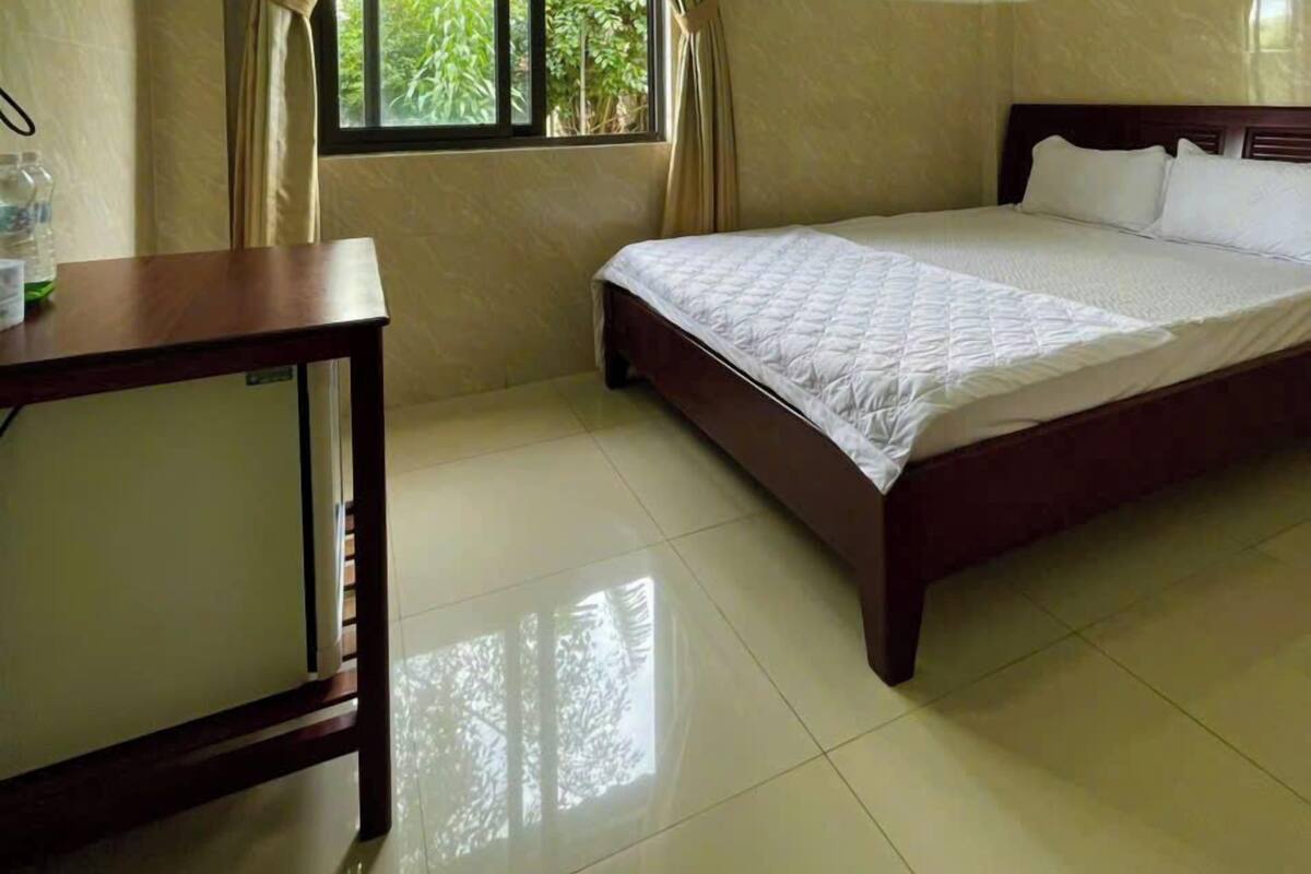 Una habitación de hotel con cama, una mesita, una ventana con cortinas y un equipo de aire acondicionado.