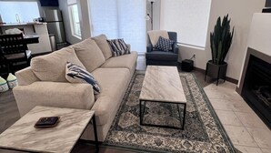 Un salon moderne avec un canapé, une table basse et un fauteuil, une cheminée, et un coin cuisine à l’arrière-plan.