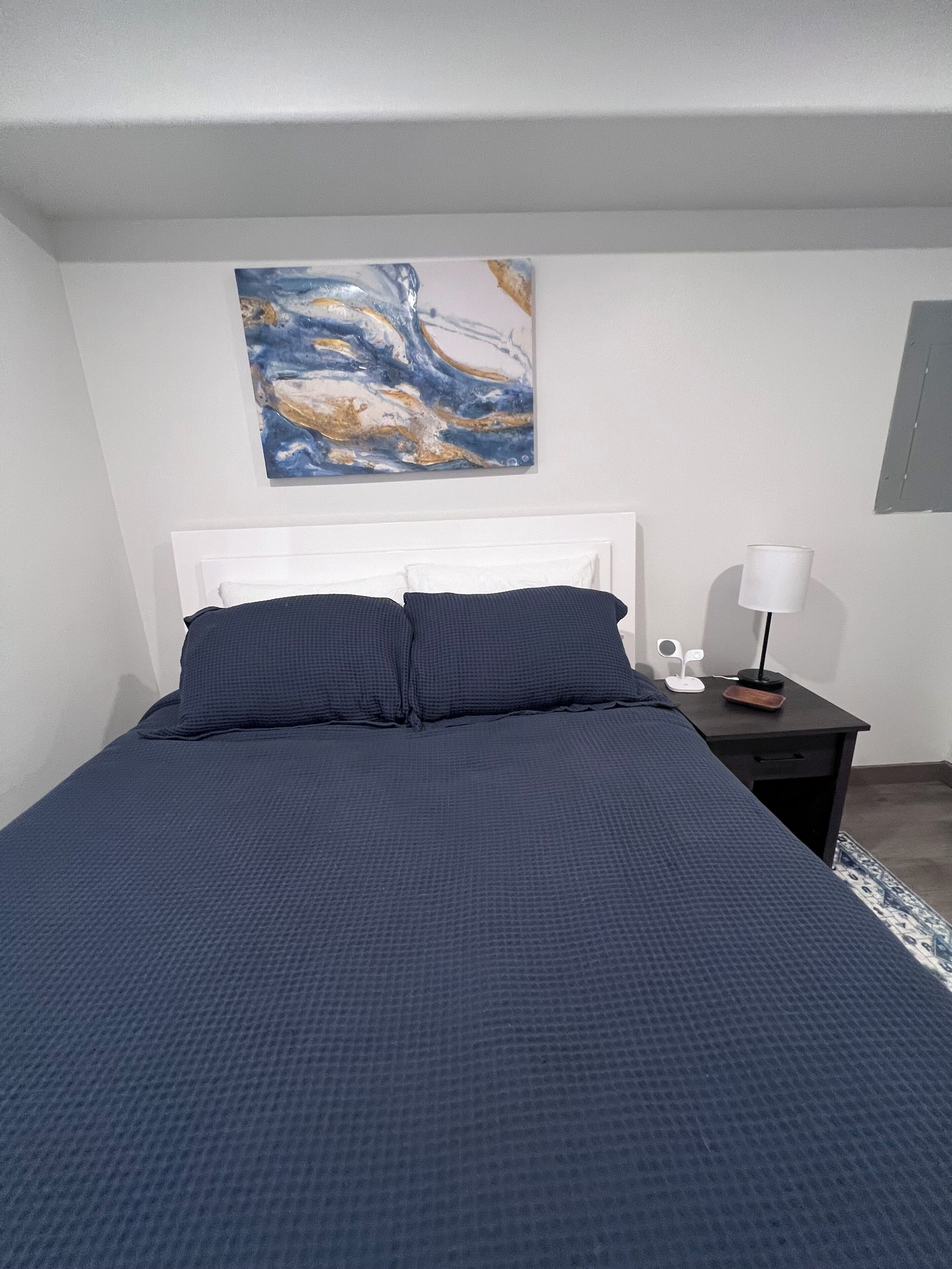 A bedroom with a bed, a nightstand, a lamp, and a painting on the wall.