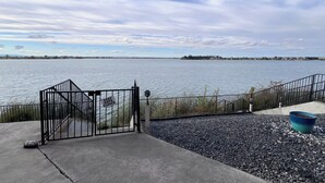 Un chemin en béton mène à une entrée fermée par un portail, qui donne sur une étendue d’eau avec une rive éloignée.