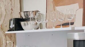A kitchen with a sink, a coffee maker, and shelves holding glasses and a bowl.