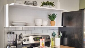 A kitchen with a black refrigerator, a stove, and a blender on the counter. There are white cabinets above the counter and a shelf with dishes and plants.