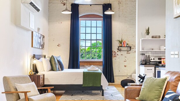 A modern bedroom with a bed, a chair, a sofa, a window with curtains, and a kitchenette.