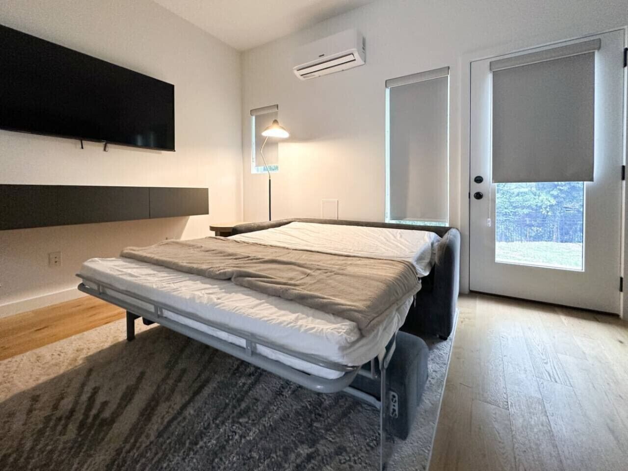Une chambre à coucher comprenant un lit, un téléviseur fixé au mur, un climatiseur et une porte donnant sur un balcon.