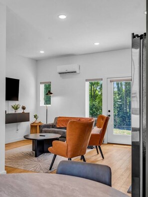 A modern living room with a sofa, chairs, a coffee table, and a TV. There are windows with a view of greenery outside. - Luxury Townhouse w/Rooftop (Knoxville)