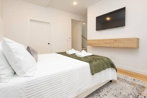 A hotel room with a bed, a flat-screen TV mounted on the wall, and a wooden shelf below it. - Luxury Townhouse w/Rooftop (Knoxville)