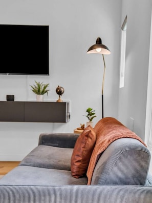A modern living room with a grey sofa, a flat-screen TV mounted on the wall, a floor lamp, and a small side table with decorative items. - Luxury Townhouse w/Rooftop (Knoxville)
