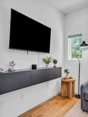 A modern living room with a large flat-screen TV mounted on the wall, a wooden sideboard with decorative items, a small round wooden table, and a floor lamp. - Luxury Townhouse w/Rooftop (Knoxville)