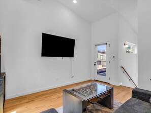 A modern living room with a flat-screen TV mounted on the wall, a glass-top coffee table, and a wooden floor. - Luxury Townhouse w/Rooftop (Knoxville)