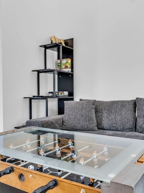 A foosball table in a modern living room with a grey sofa and a black shelving unit in the background.