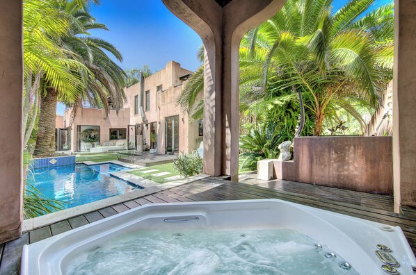 A hot tub in a courtyard with a pool, surrounded by palm trees and a building with large windows.