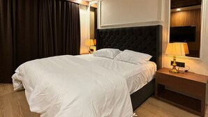 A bedroom with a large bed, wooden floor, a dark headboard, a nightstand with a lamp, and a window with curtains. - The Magnolia Srinagar (Srinagar)