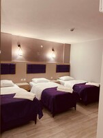 A hotel room with four beds, each with a purple bedspread and white linens. There are wall-mounted light fixtures above each bed.