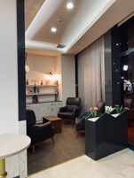 A modern living room with a marble floor, leather sofas, a coffee table, and a fireplace. There is a bookshelf with decorative items and a large window with curtains.