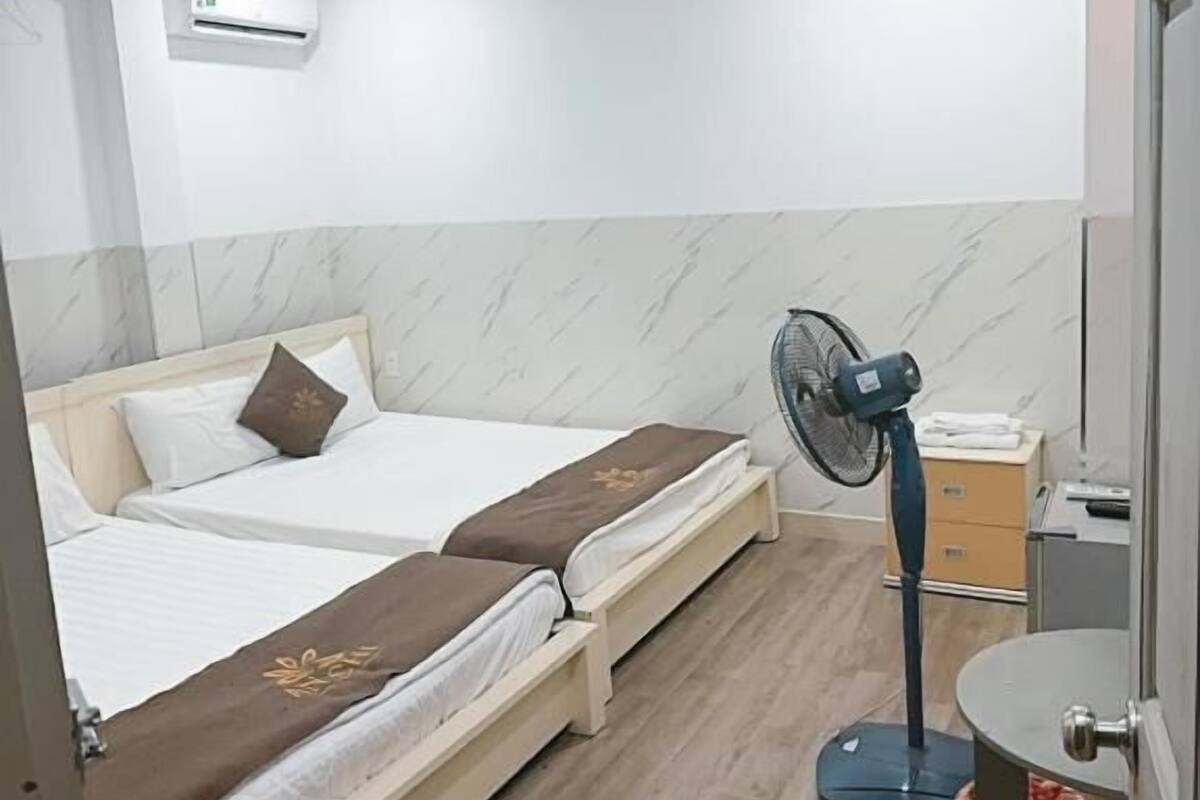 A hotel room with two single beds, a standing fan, a small table with a red basket, and a wall-mounted air conditioner.