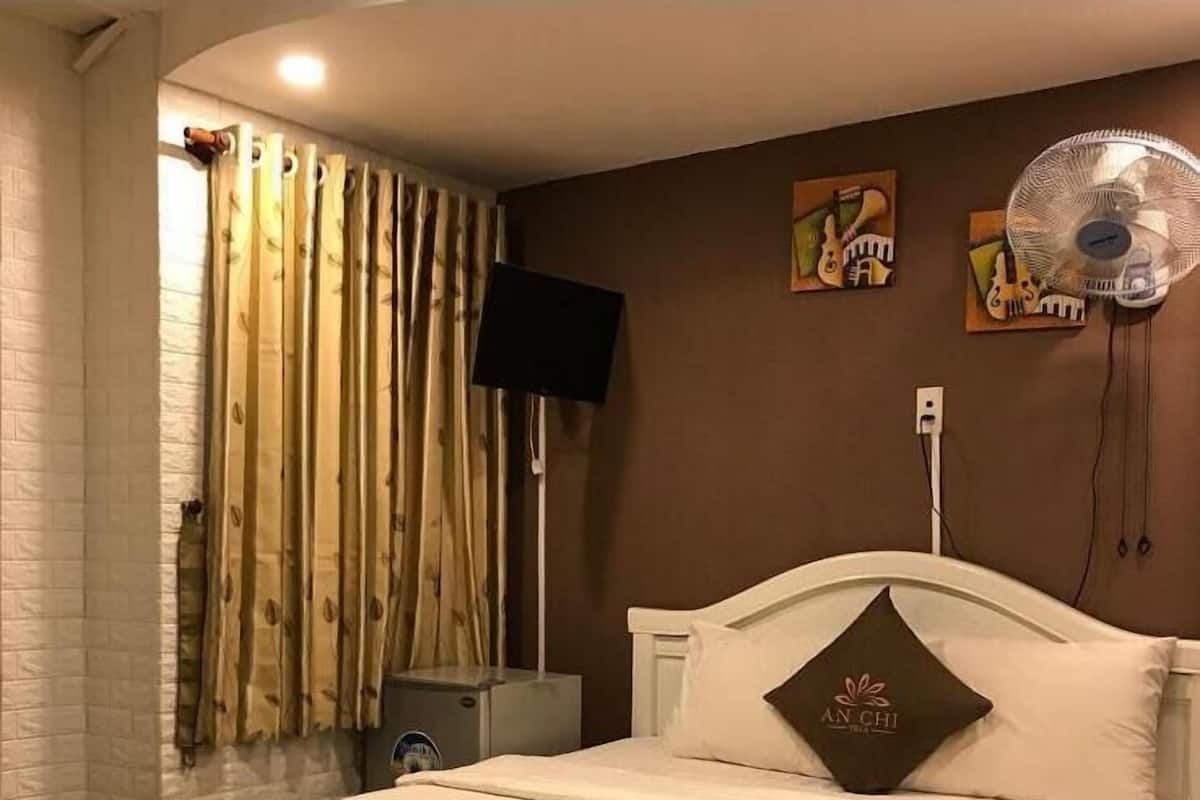 A neatly made bed with a brown bedspread and a pillow with a logo, a wall-mounted fan, and a painting on the wall.