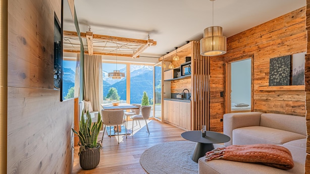 Een moderne woonkamer met houten wanden, een bank, een eethoek met tafel en stoelen, en uitzicht op bergen door grote ramen.