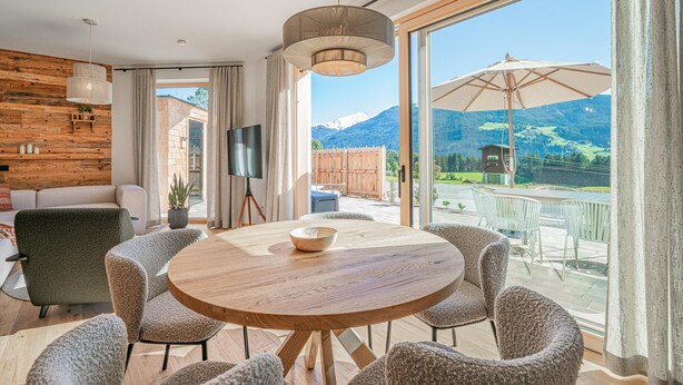 Uma sala de estar moderna com uma mesa redonda de madeira, assentos confortáveis, uma TV sobre um tripé e uma janela grande com vista para as montanhas.