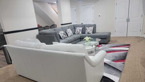 Een moderne woonkamer met een grijze hoekbank, een witte bank en een glazen salontafel. Op de vloer ligt een kamerkleed met een patroon.
