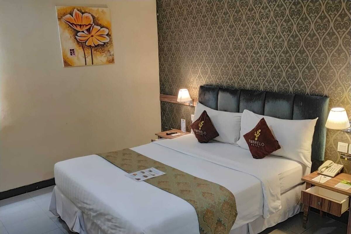 A hotel room with a bed, a nightstand, a lamp, and a wall with a patterned wallpaper.