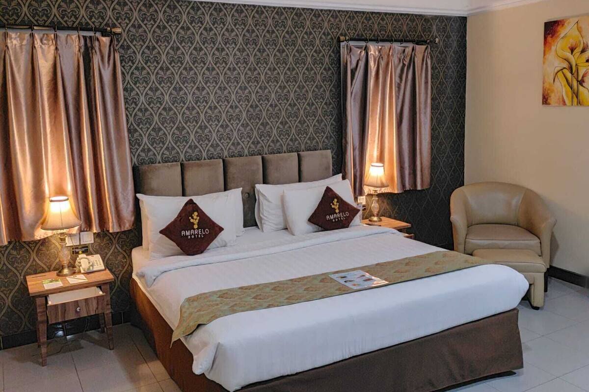 A hotel room with a large bed, two bedside lamps, a chair, and a patterned wallpaper.