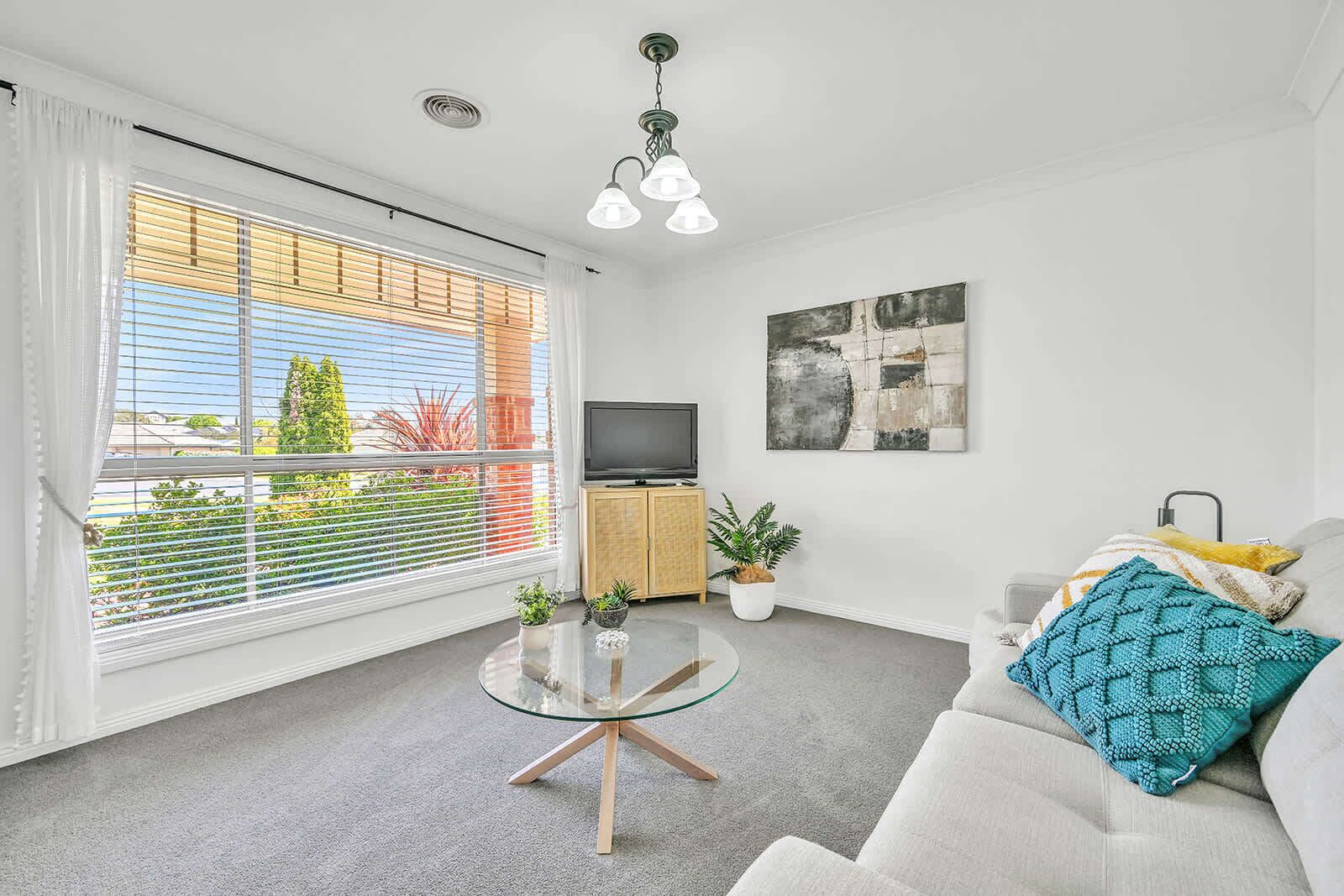 Een moderne woonkamer met een glazen salontafel, een bank met een decoratief plaid, een flatscreen televisie en een groot raam met zonwering.
