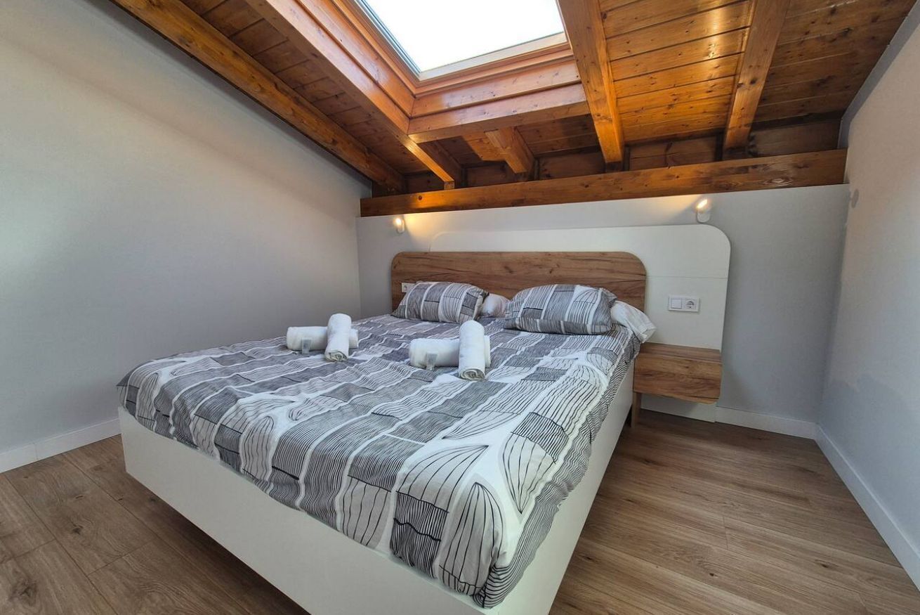 Une chambre avec un puits de lumière, des poutres en bois au plafond, un lit recouvert d’une courtepointe à rayures et une tête de lit blanche.