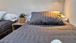 Een eenpersoonsbed met een gestreepte deken, een nachtkastje met een geweven mandje en een kleine plant, en een wandlamp.
