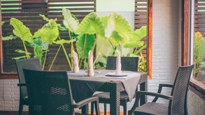 A table set for four with wicker chairs, a white tablecloth, and large green plants in the background.
