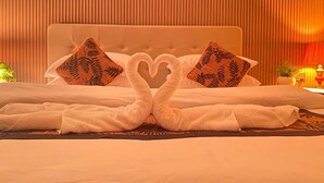 A hotel room with a bed featuring two pillows with a leaf pattern, a heart-shaped towel arrangement, and a warm ambient light.