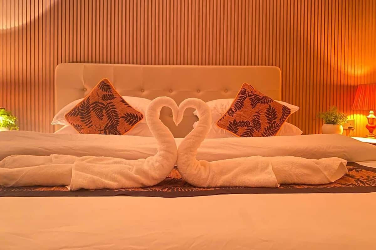 A hotel room with a bed featuring two pillows with a leaf pattern, a heart-shaped towel arrangement, and a warm ambient light.